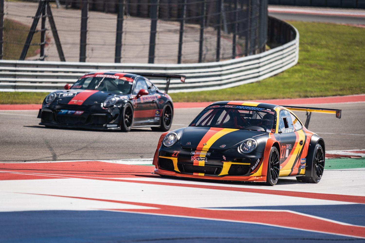 Two Porsche GT race cars battle for position through a corner on a professional circuit.