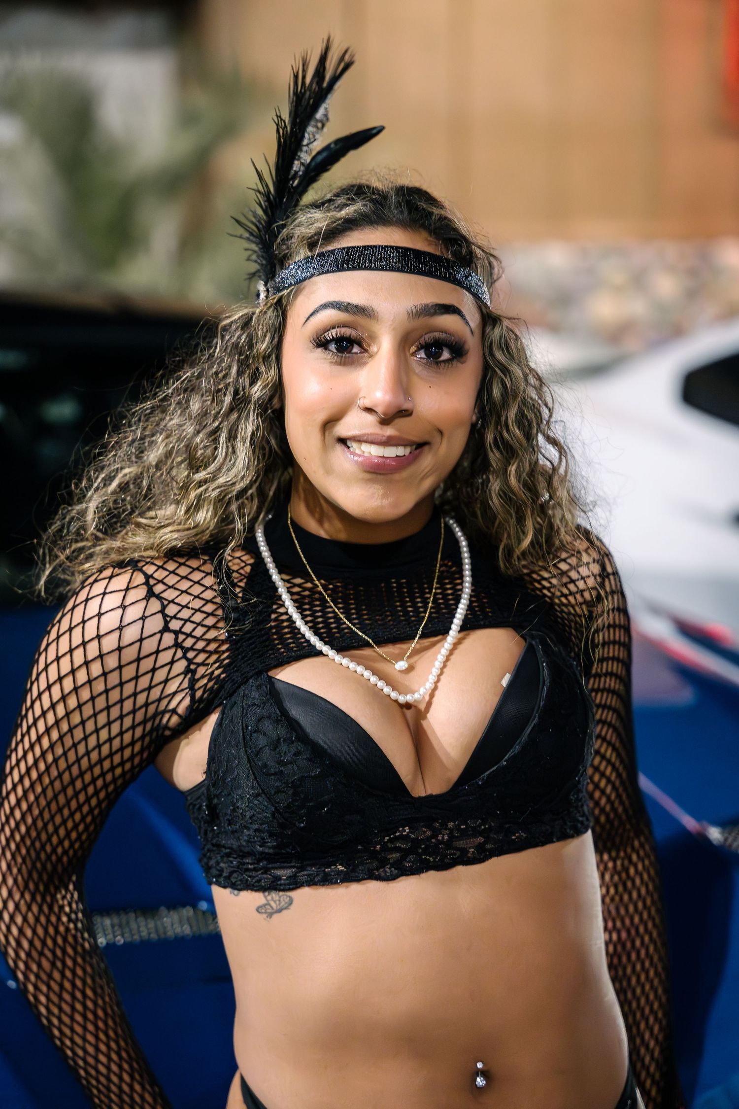 Model in a black fishnet outfit with feather headband poses at a car event.