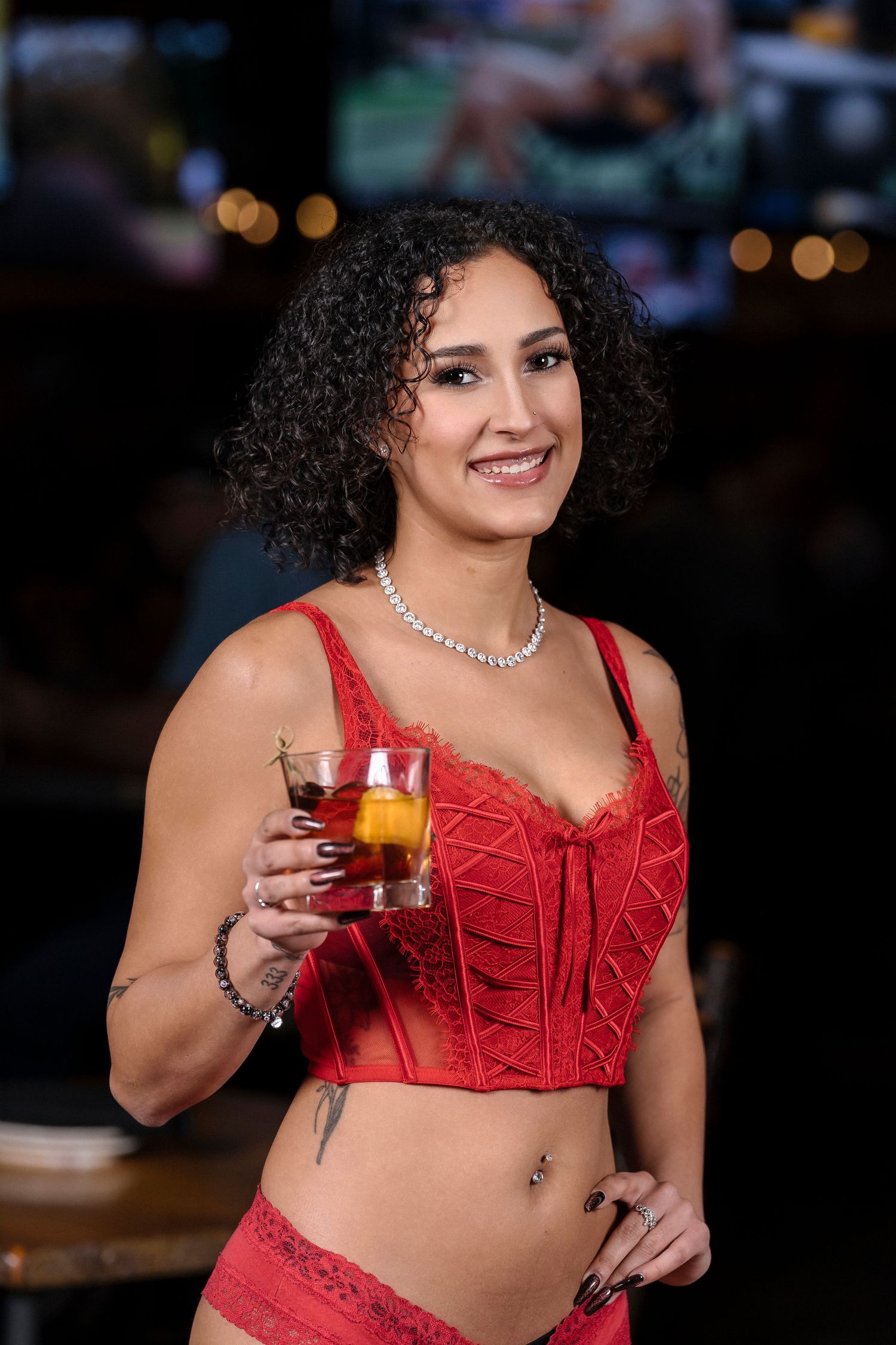 Woman in red lace lingerie holds a cocktail while posing at a bar.