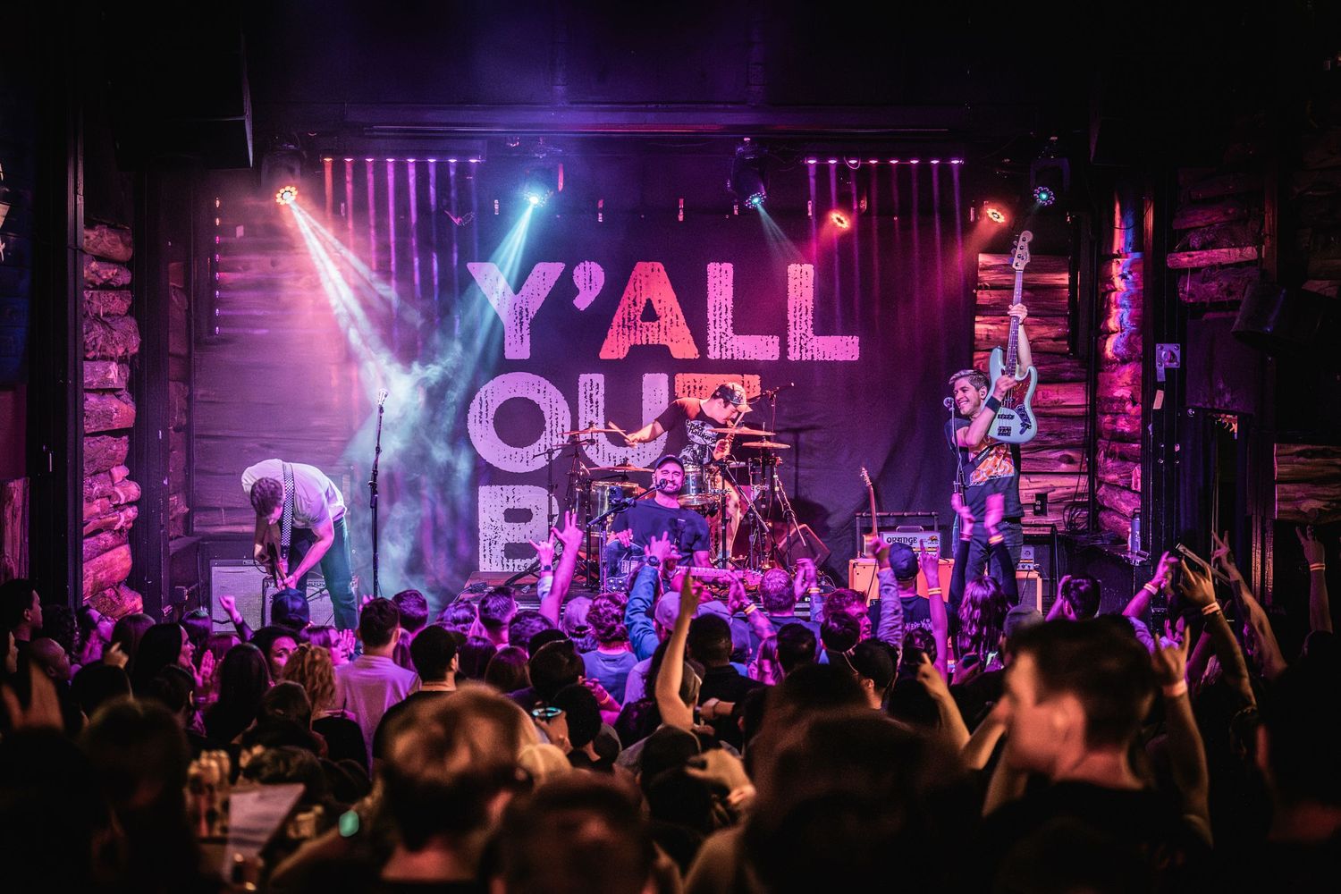 Enthusiastic concert audience with raised hands watching a band perform under colorful stage lights.