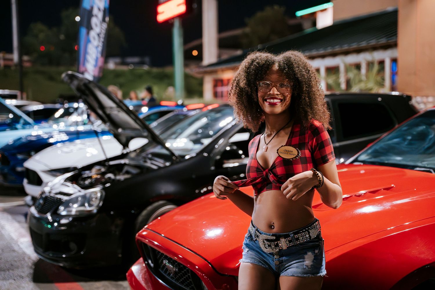 Model in casual red plaid outfit poses next to red car at nighttime car meet.