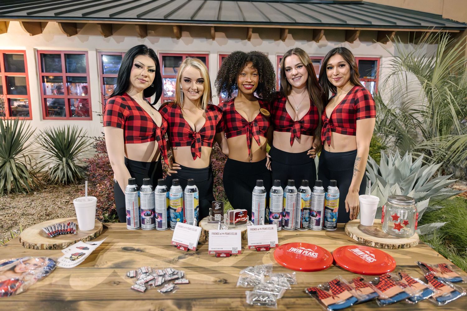 Group in matching red plaid crop tops posing with beverages and snacks on wooden display table.