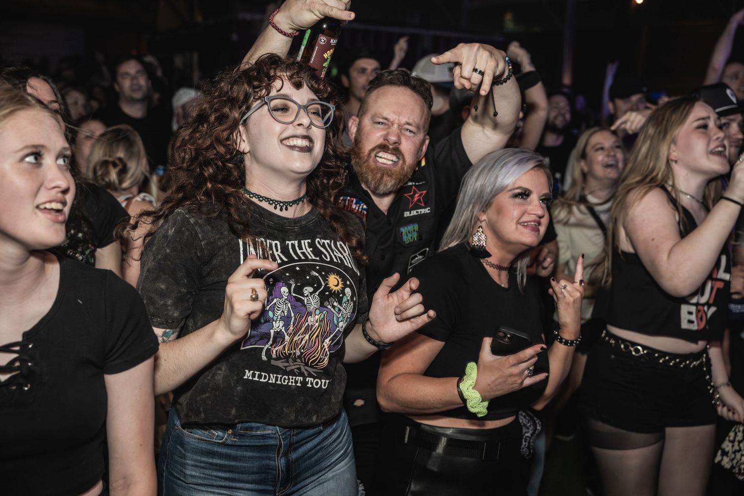 An energetic crowd enjoys a concert with fans wearing band t-shirts and throwing up rock hand gestures.