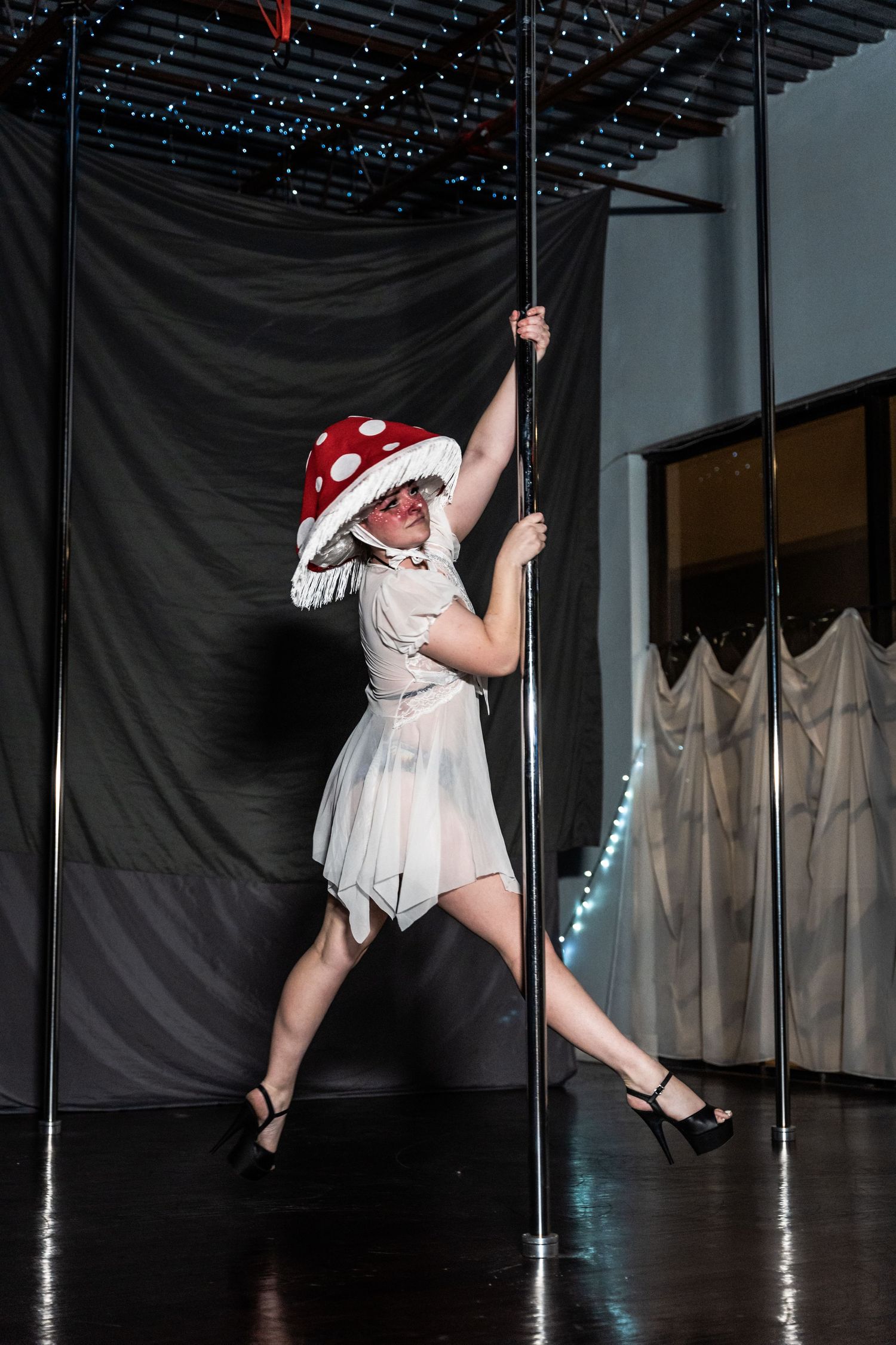 Performer in white dress and polka dot hat doing pole dance routine with twinkle lights overhead.