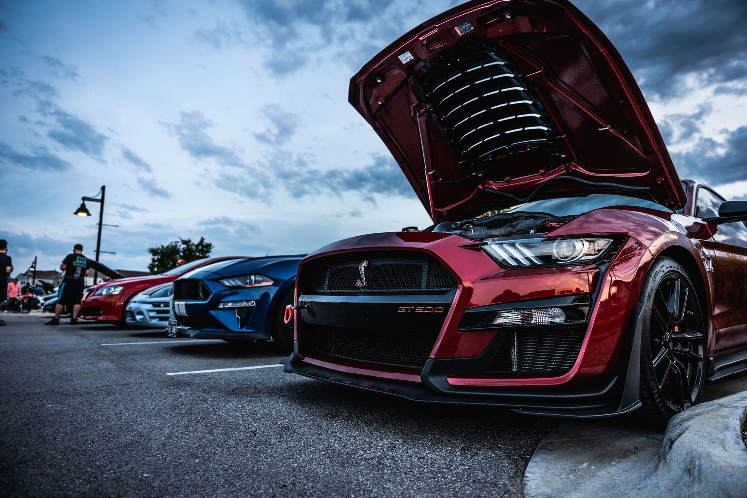 Modified red Audi GT with open hood showcasing engine bay at a car meet under dramatic cloudy sky.
