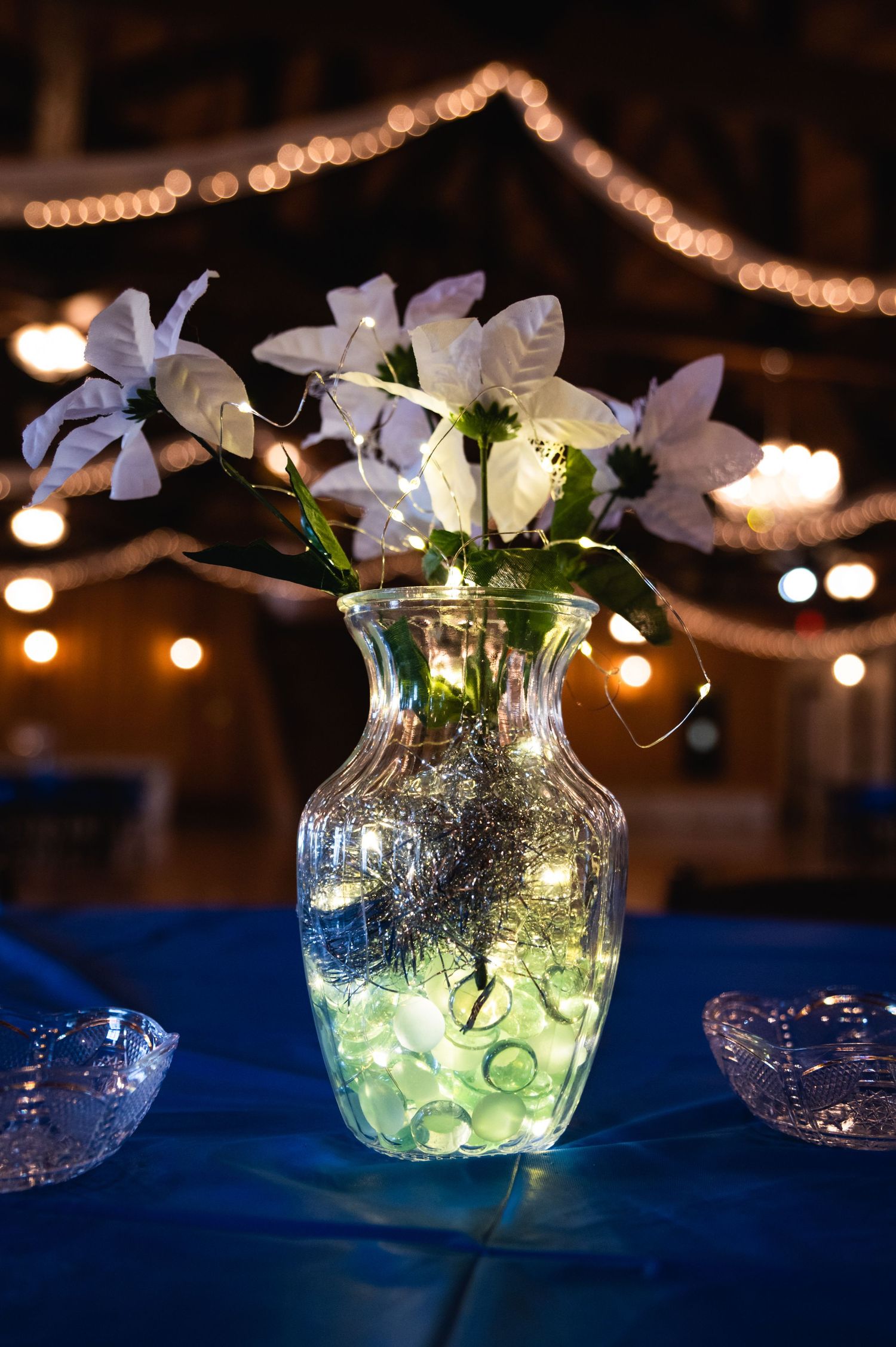 Illuminated floral centerpiece with white orchids in a glass vase against twinkling reception lights.