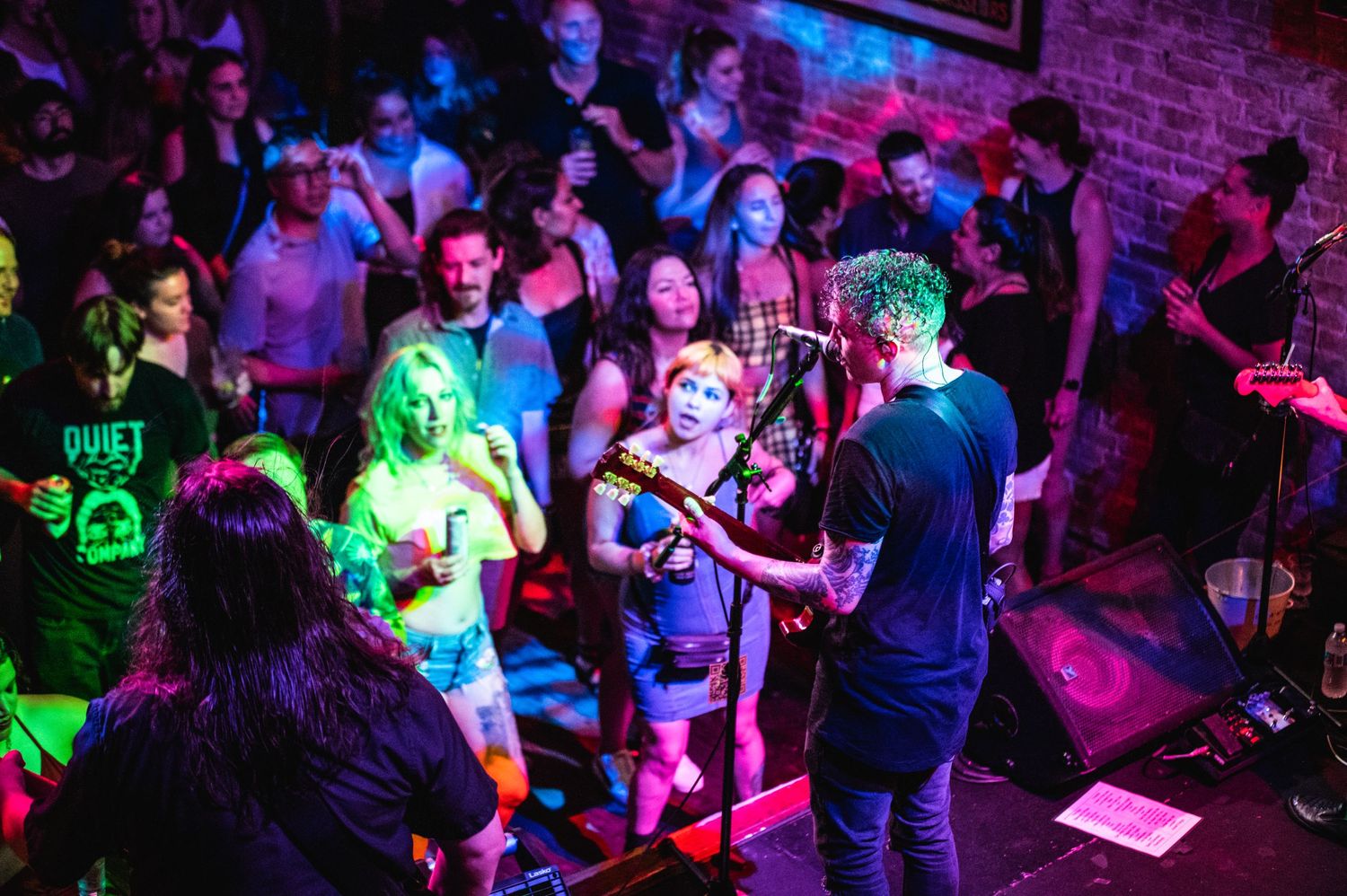 Concert crowd watches performers on stage in an intimate venue with colorful lighting effects.