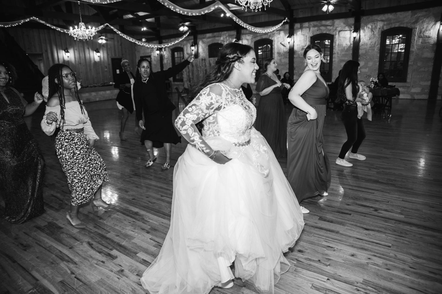 A black and white photo shows wedding guests dancing on a wooden floor under elegant draping and chandeliers.