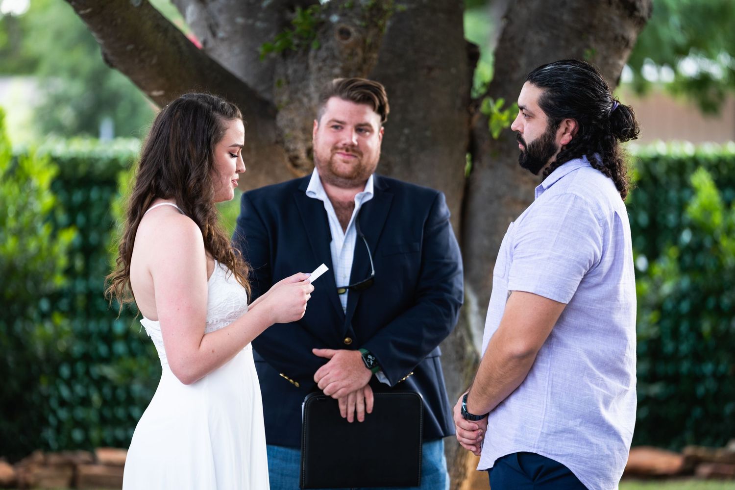 Three people having a conversation during an outdoor ceremony under a large tree.