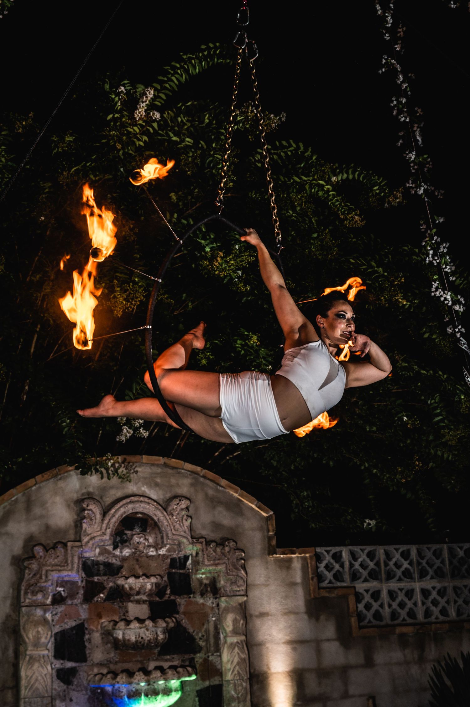 Aerial performer in white costume executes dynamic pose surrounded by fire elements at night.