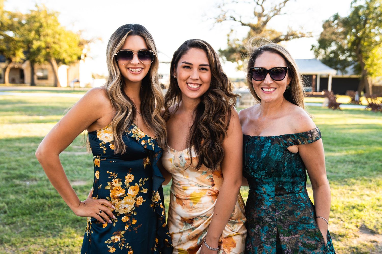 Three women in floral dresses pose together at an outdoor event.