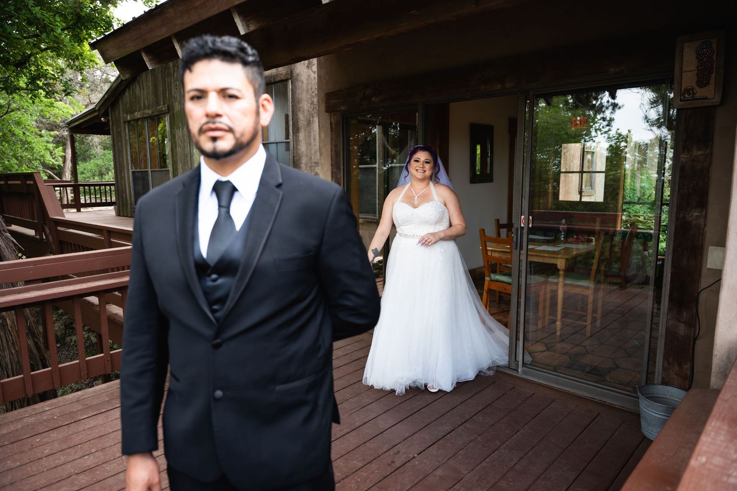A wedding couple stands on a rustic wooden deck with the bride in white and groom in black suit.