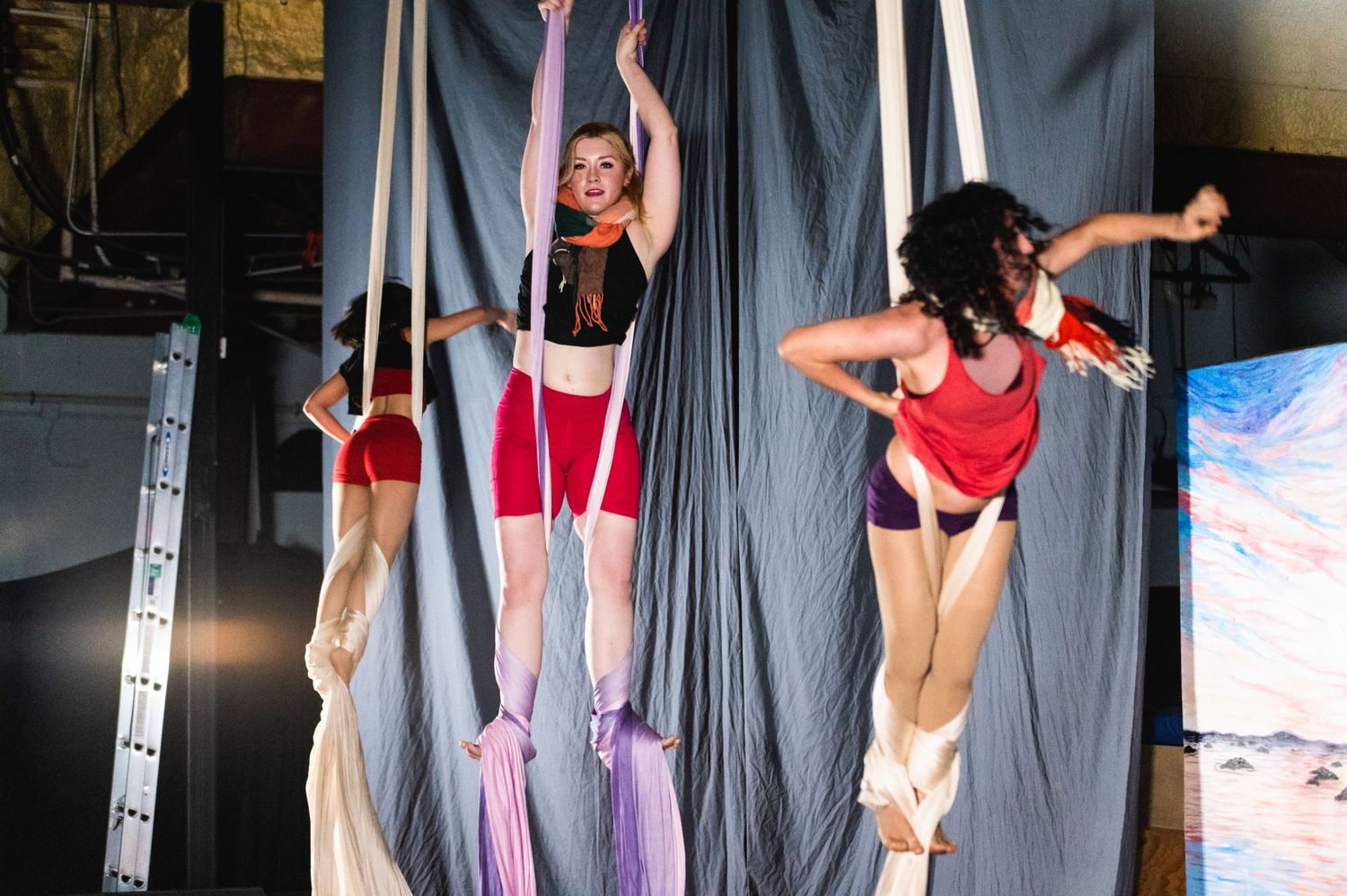Three aerial performers execute synchronized moves on suspended silks against a grey backdrop.