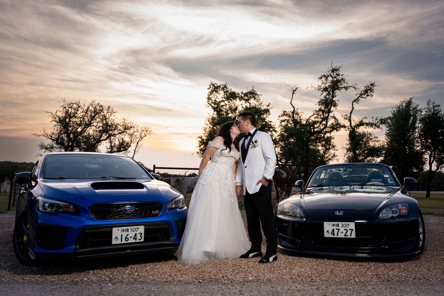 Japanese sports cars Subaru WRX STI and Honda S2000 parked at sunset with newlyweds posing between them.