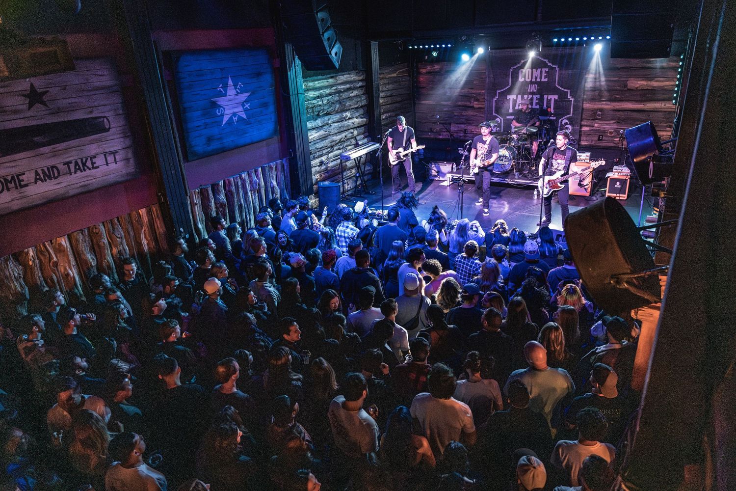 Crowded music venue with stage and blue lighting showing band performing under lone star flag.