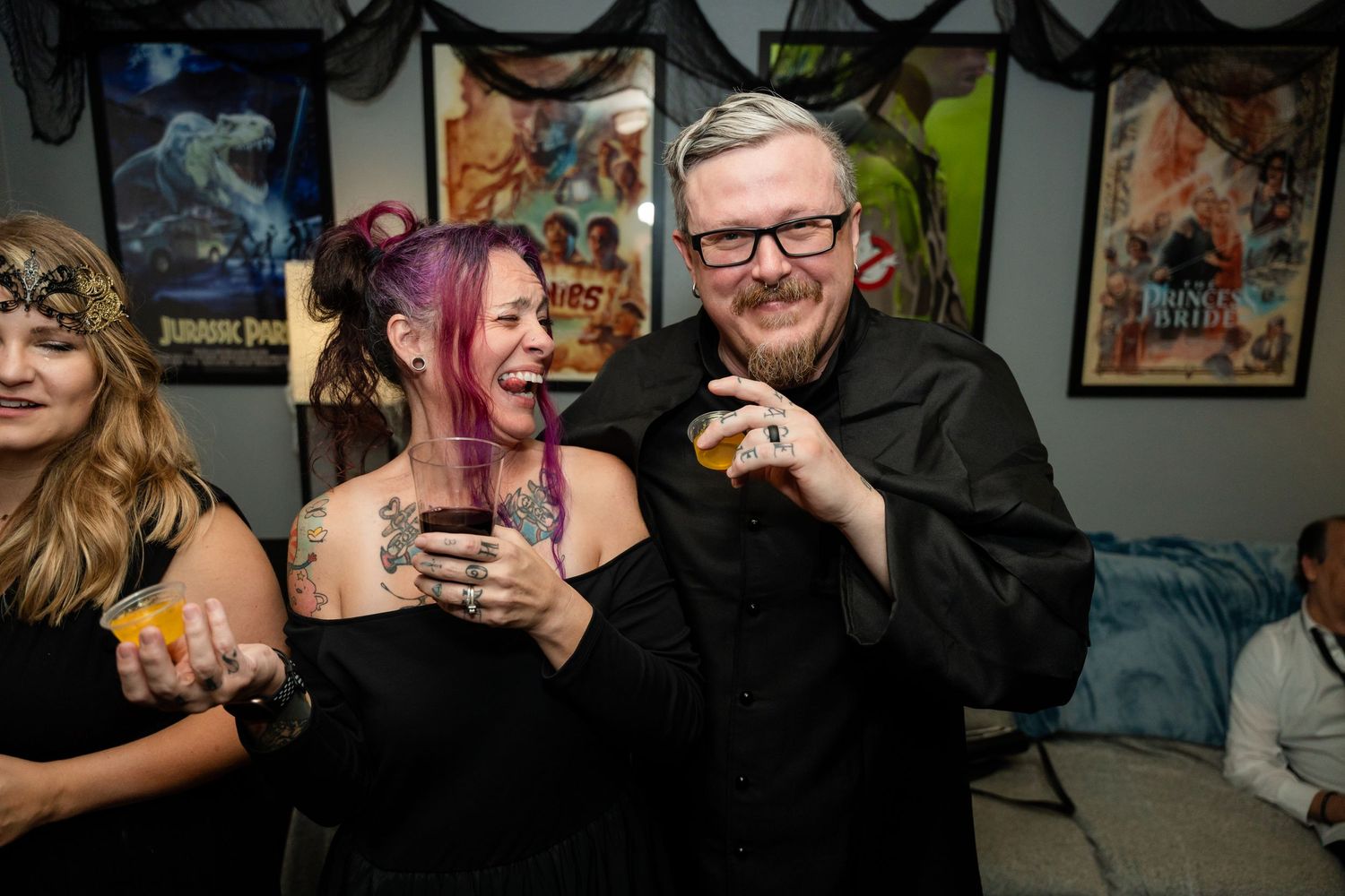 Partygoers enjoying drinks and laughing at indoor celebration with movie posters on wall.