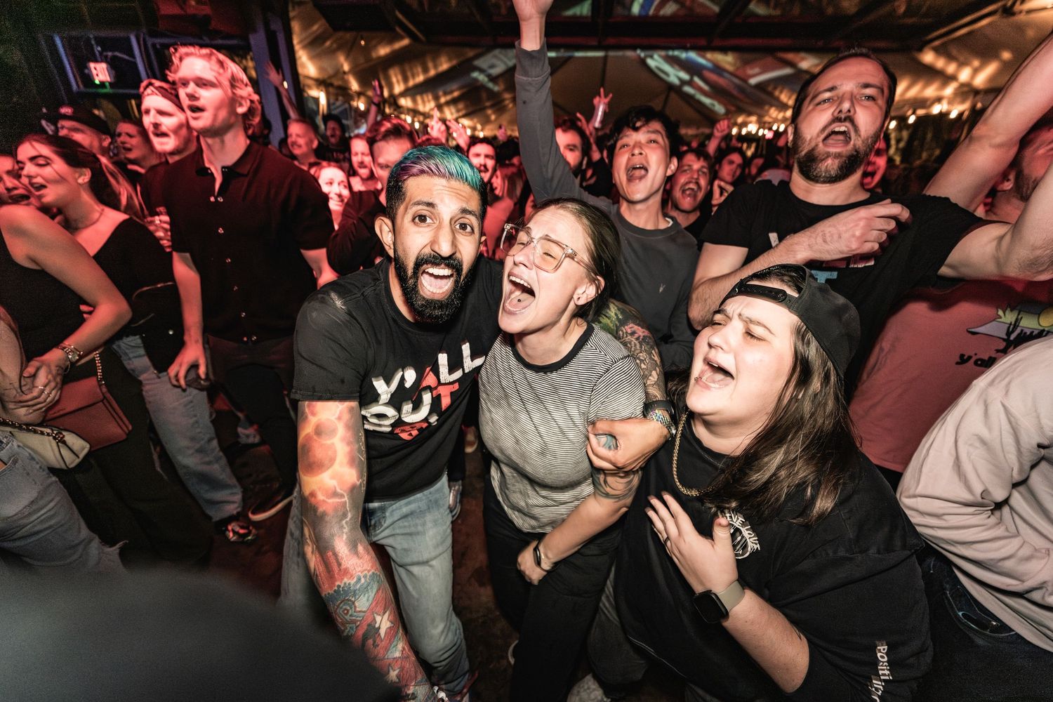 An enthusiastic crowd of concert-goers reacts energetically during a live music performance at a packed venue.