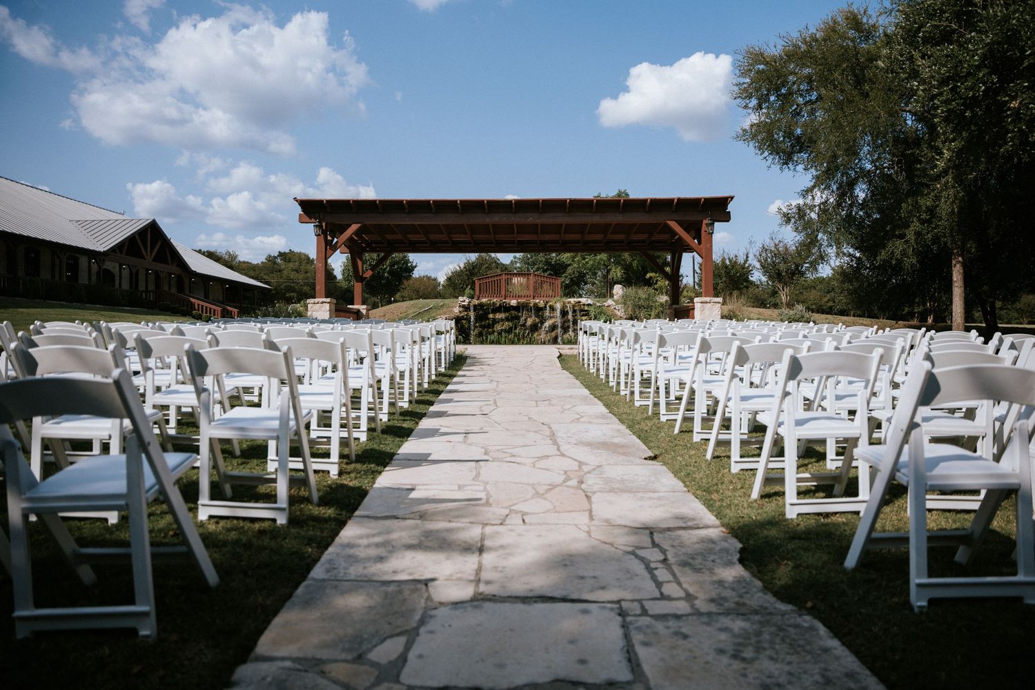 Outdoor wedding ceremony setup with white chairs leading to wooden pergola.