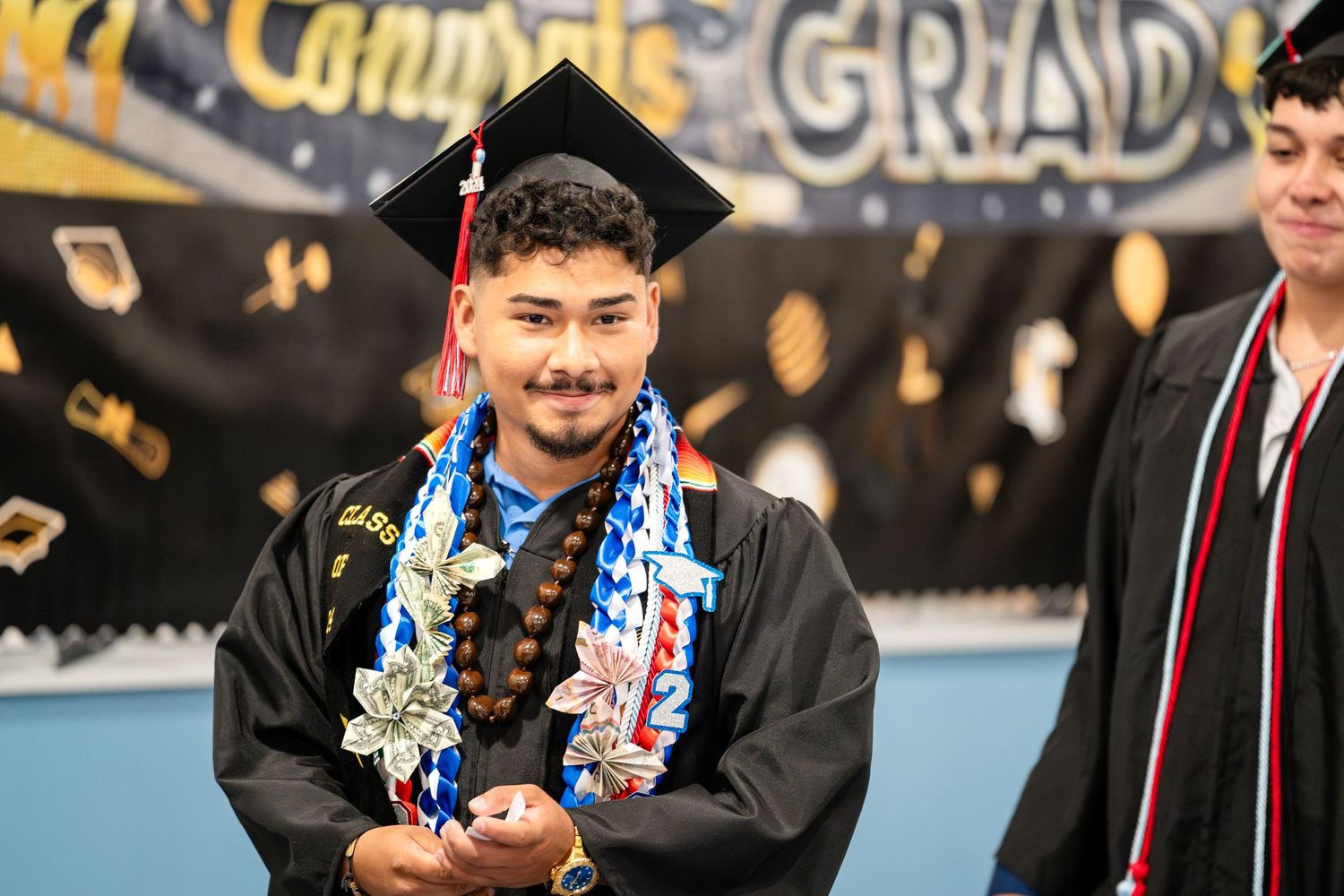 Graduate wearing black cap and gown with patriotic leis at commencement ceremony.