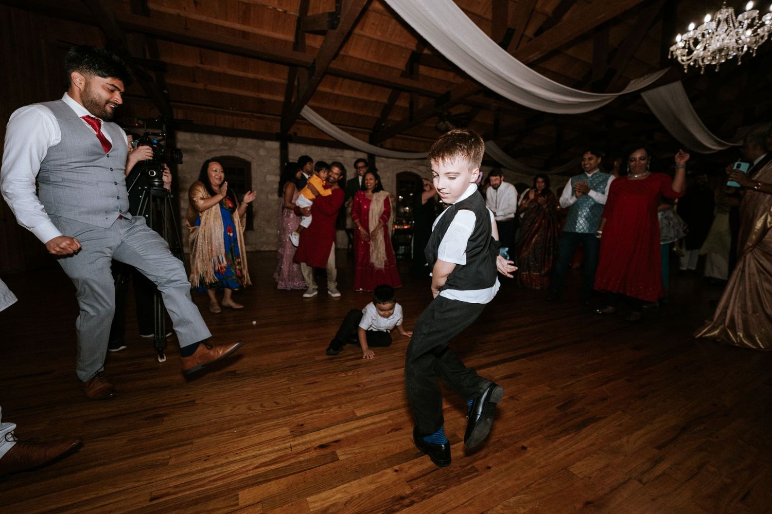 Dancing guests enjoy themselves on a wooden dance floor under elegant white draping and chandeliers.