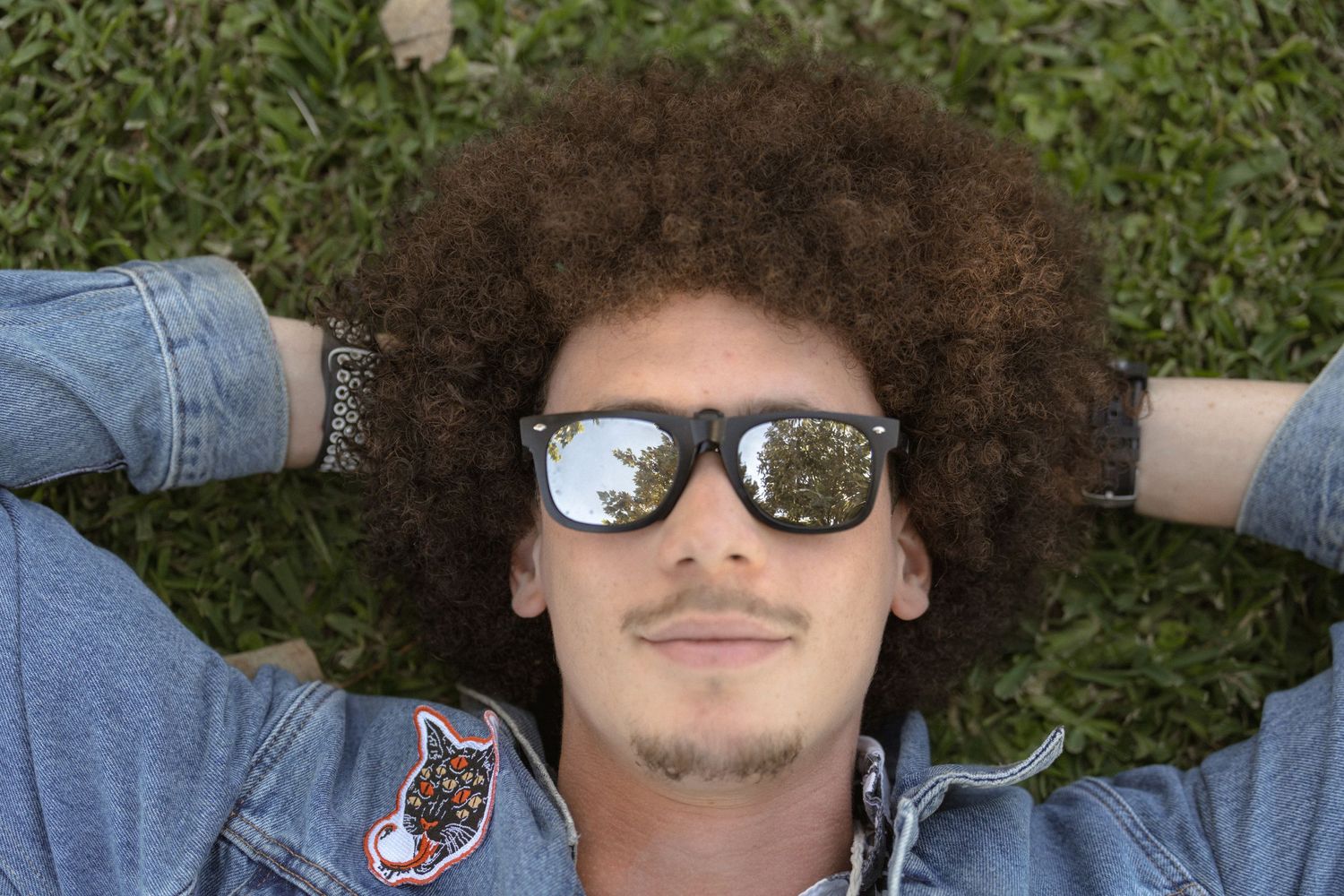 Person with large curly afro hairstyle lying on grass wearing sunglasses and denim jacket.