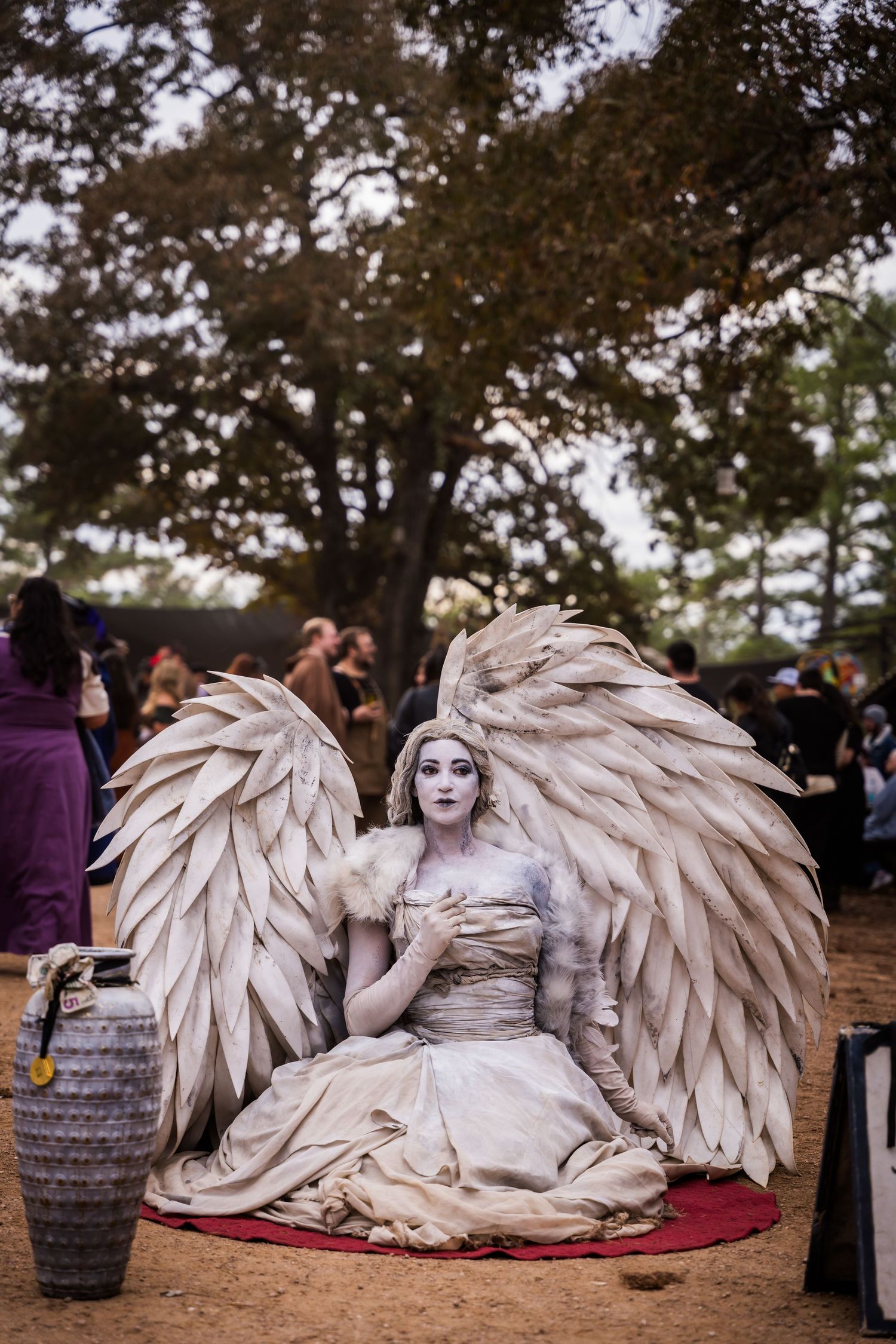 Performer dressed as an angel statue in white with large wings sits on display at an outdoor festival.