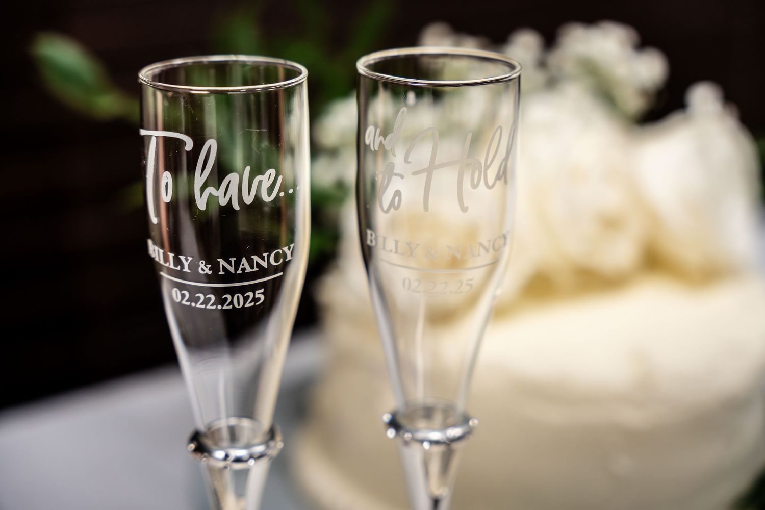 Two personalized wedding champagne flutes with engraved names and date sit elegantly against a soft blurred background.