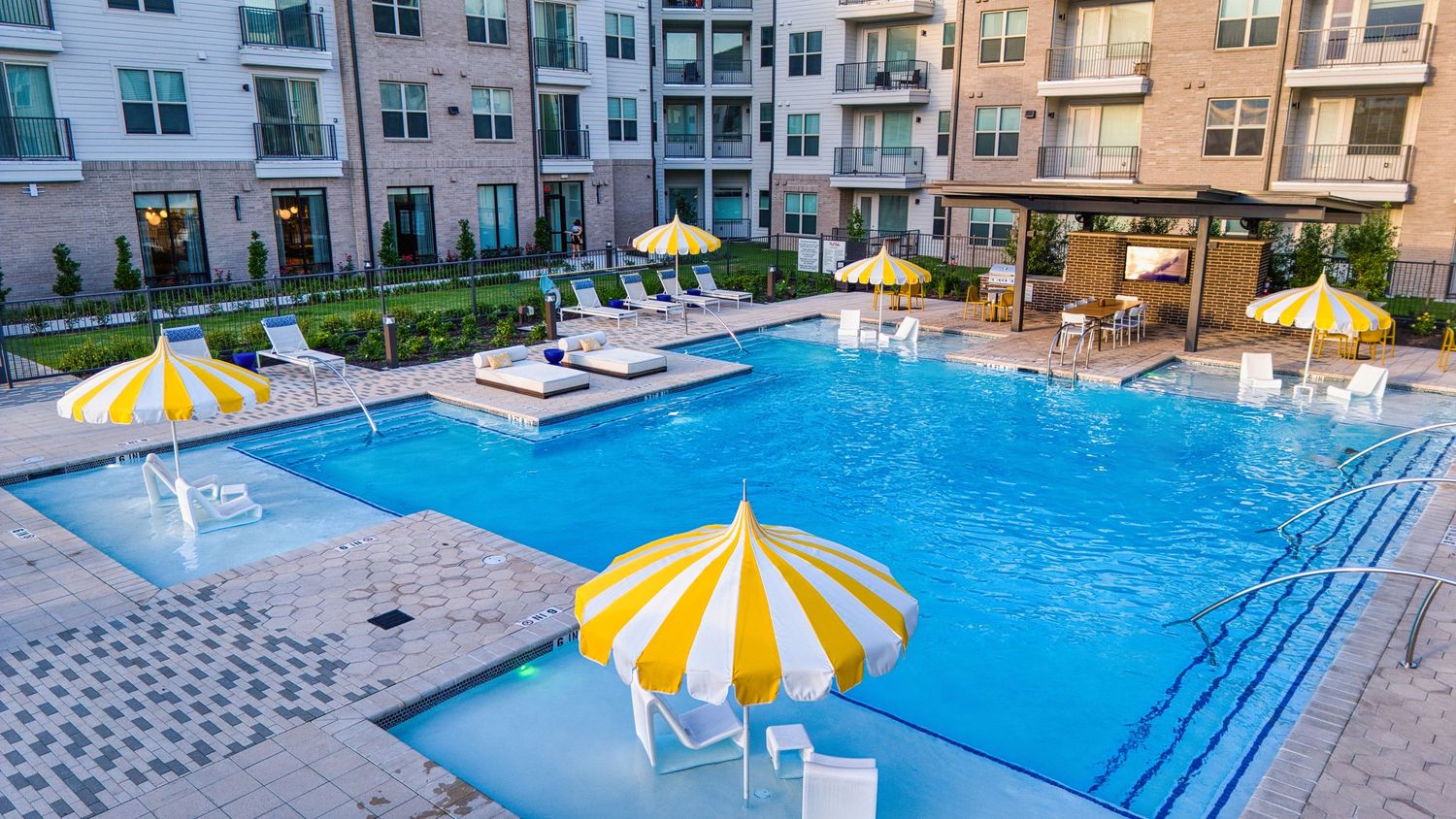 Bright blue swimming pool with striped umbrellas and lounge chairs at modern apartment complex.