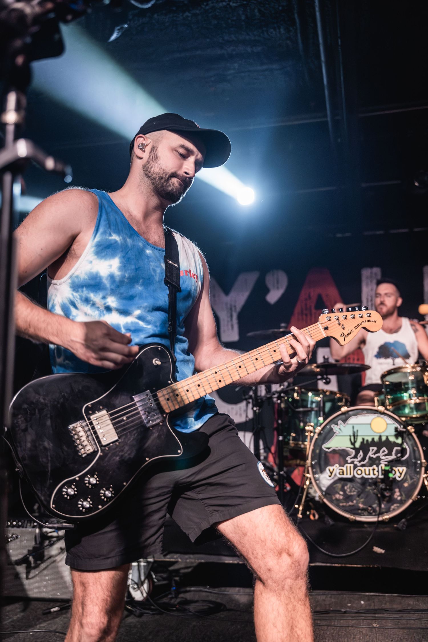 Guitarist in a tie-dye tank top performs energetically on stage with a black electric guitar.