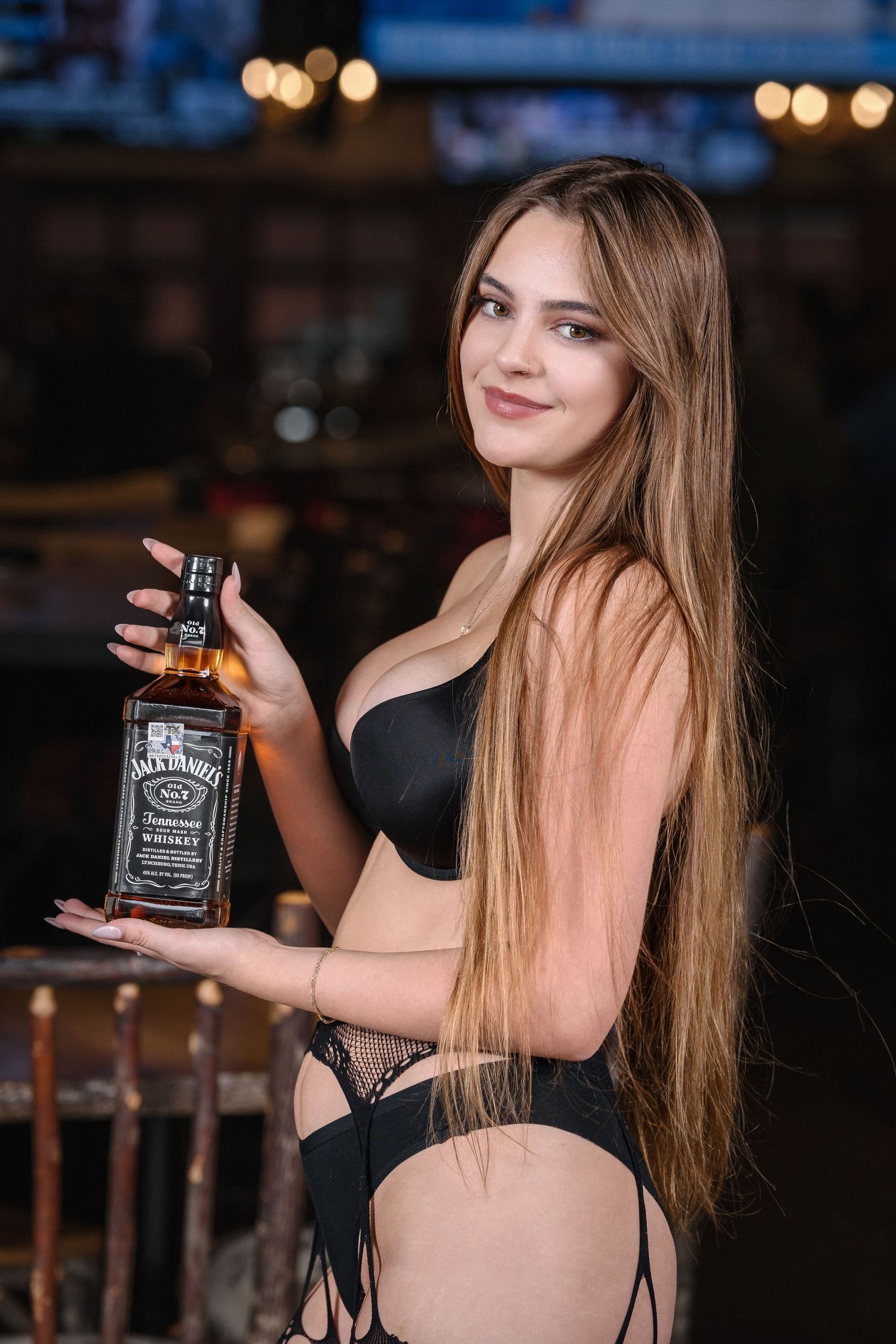Model in black lingerie holding bottle of Jack Daniels whiskey in bar setting.