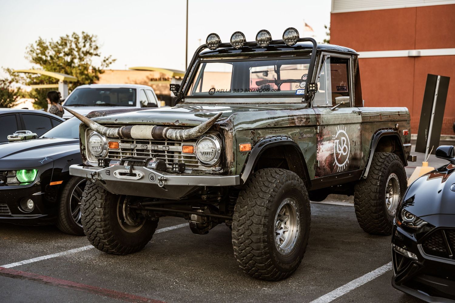 A lifted Ford Bronco with oversized tires and roof-mounted spotlights parked in a lot at sunset.