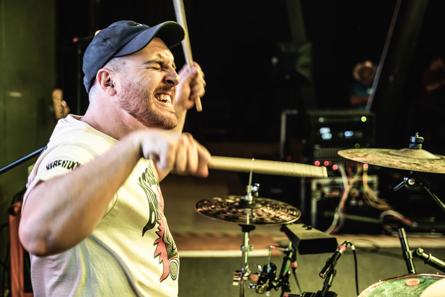 Drummer performing passionately on stage wearing a baseball cap during a concert.