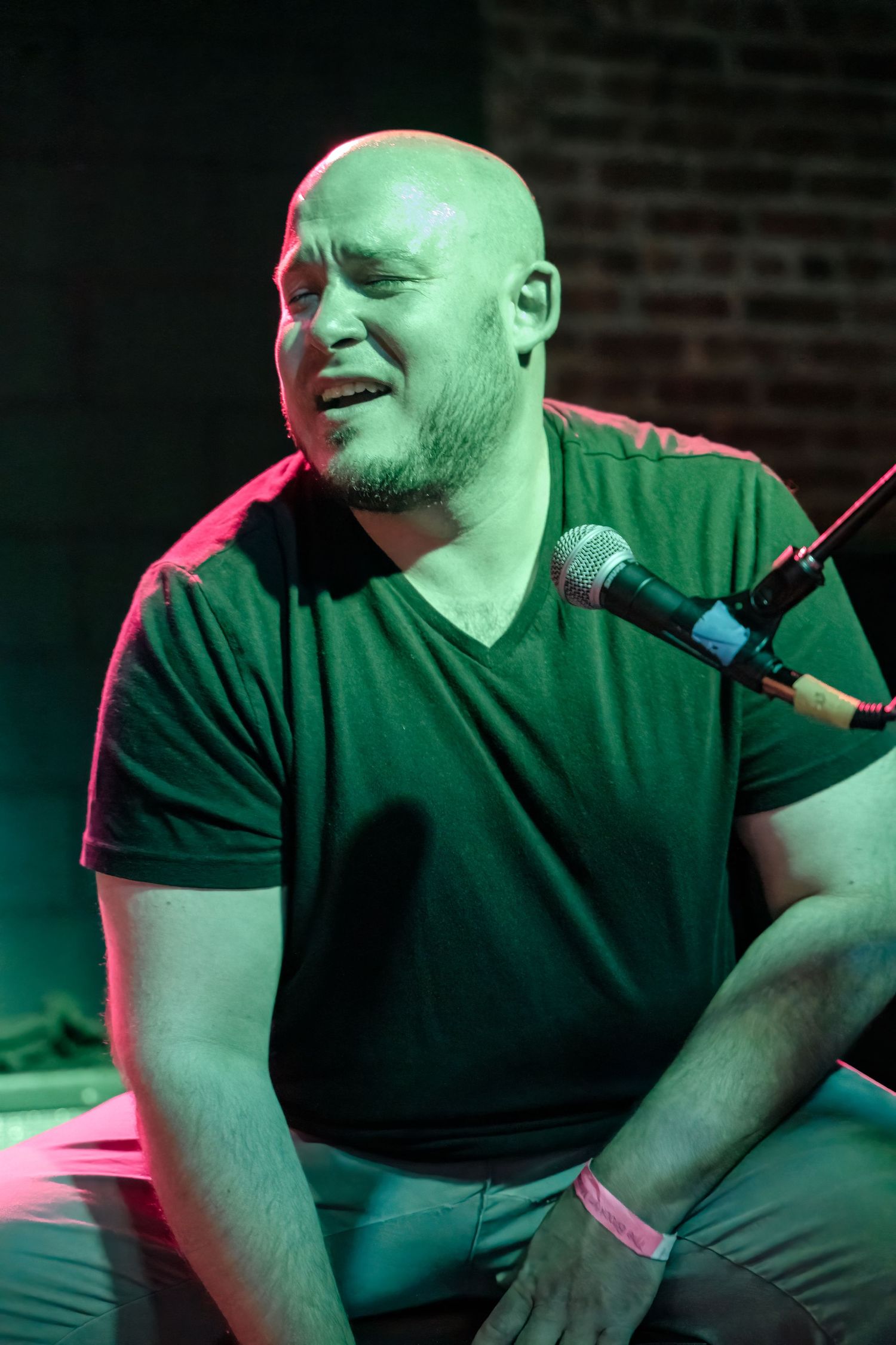 A performer in a green v-neck shirt sings emotively into a microphone on stage under dramatic lighting.