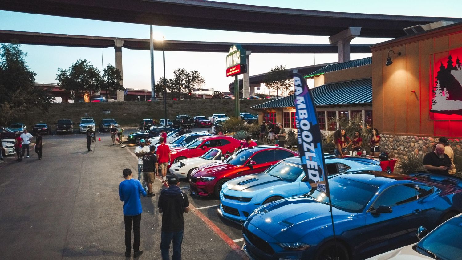Car enthusiasts gathering around modified vehicles at dusk under an elevated roadway.