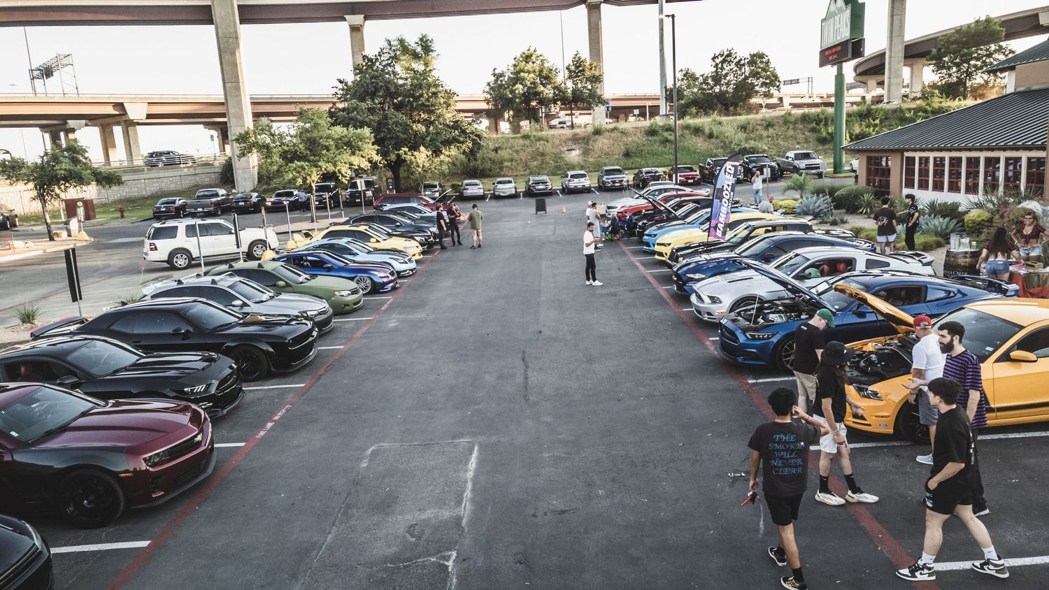 Wide angle view of car enthusiasts gathering around vehicles at outdoor car meet.
