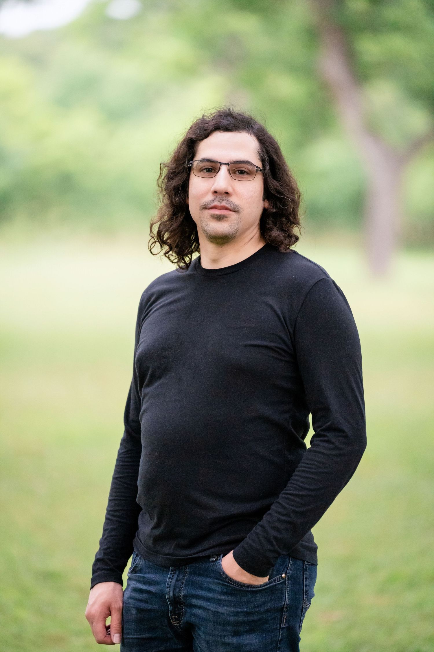 Outdoor portrait of a person in black long sleeve shirt and jeans against blurred green background.
