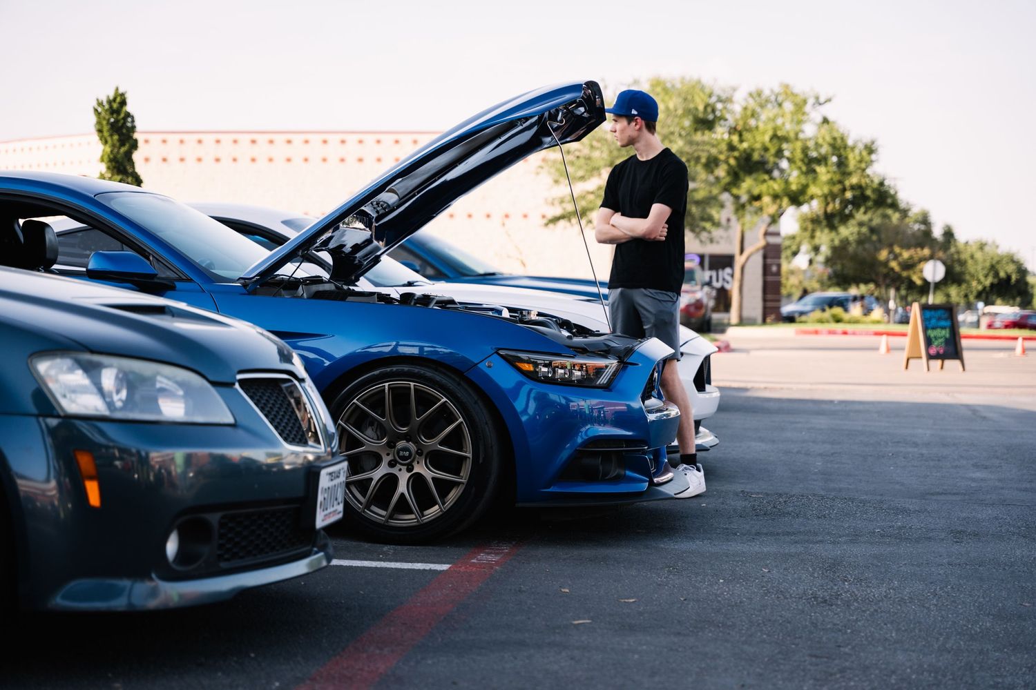 Person examining under the hood of a blue sports car in a parking lot.