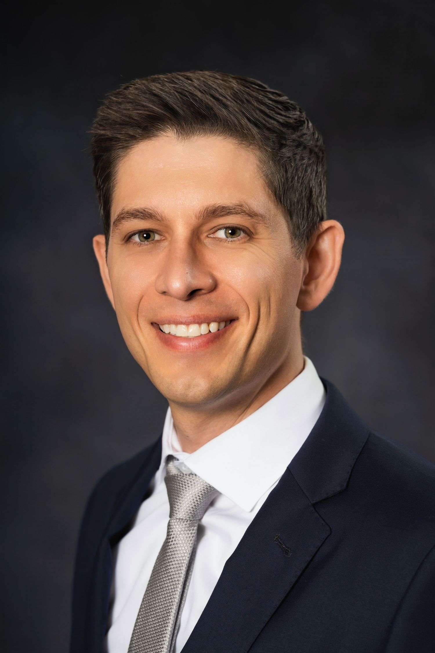 Professional headshot of person in navy suit and gray tie against dark background.