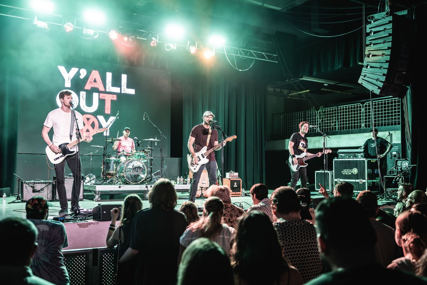 Rock band performs on stage with 'Y'all Out' banner at indoor music venue.