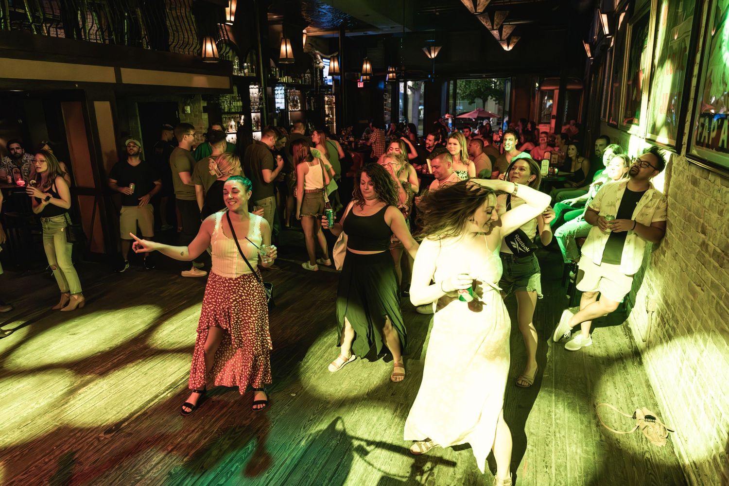 People dance together on wooden dance floor under colorful venue lighting.