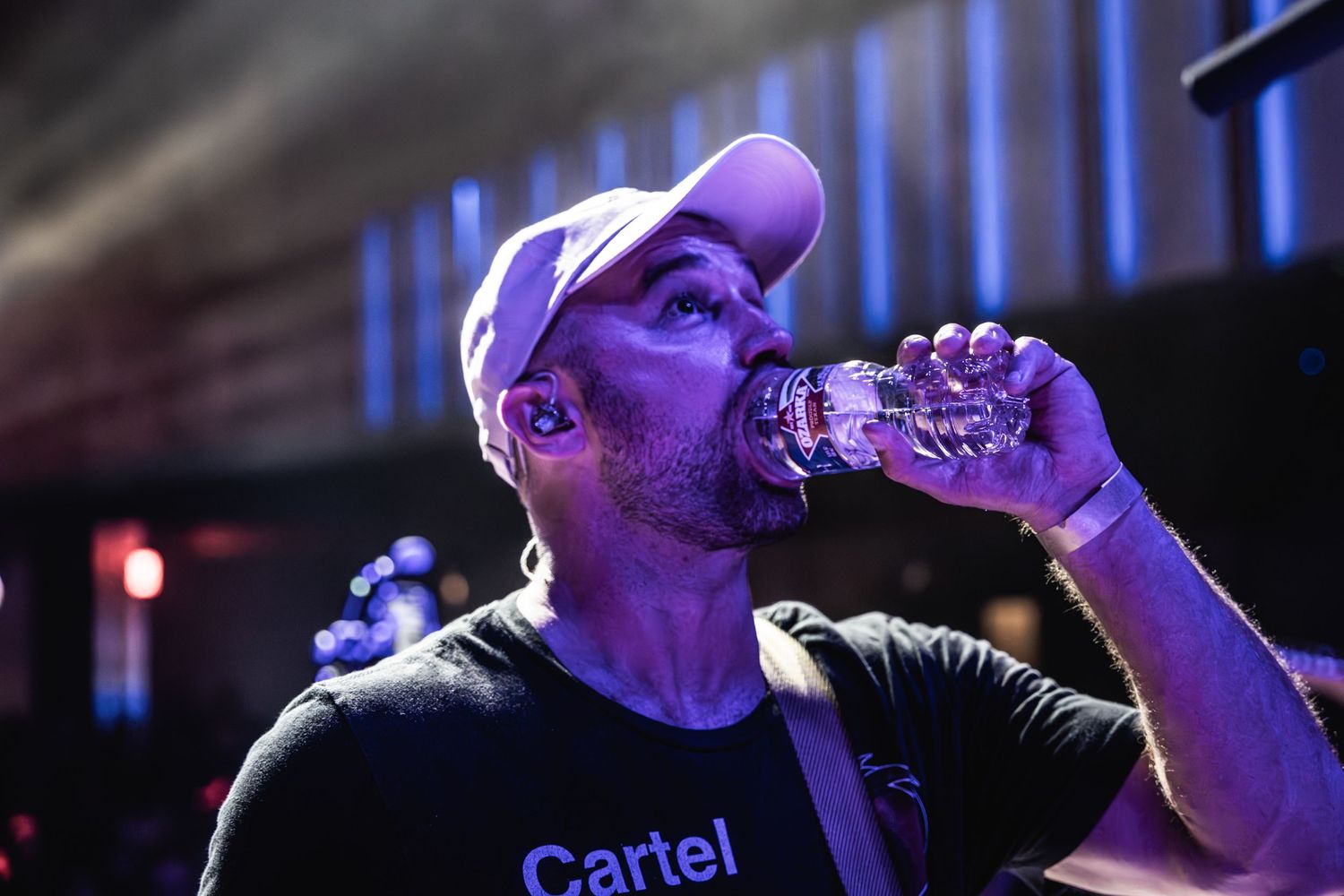 Concert performer wearing Cartel t-shirt drinking from water bottle during live show in dark venue.