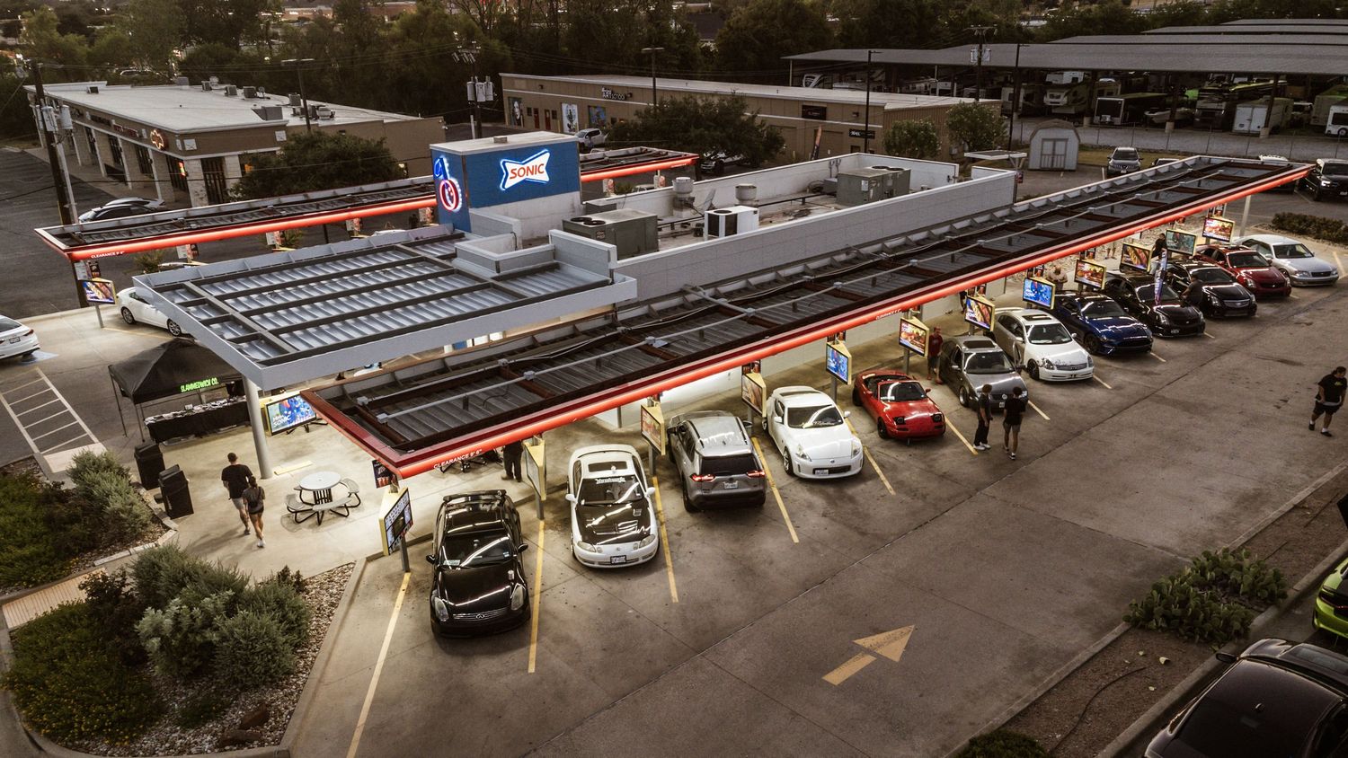 Drone shot of cars lined up at an evening automotive gathering with illuminated parking area.
