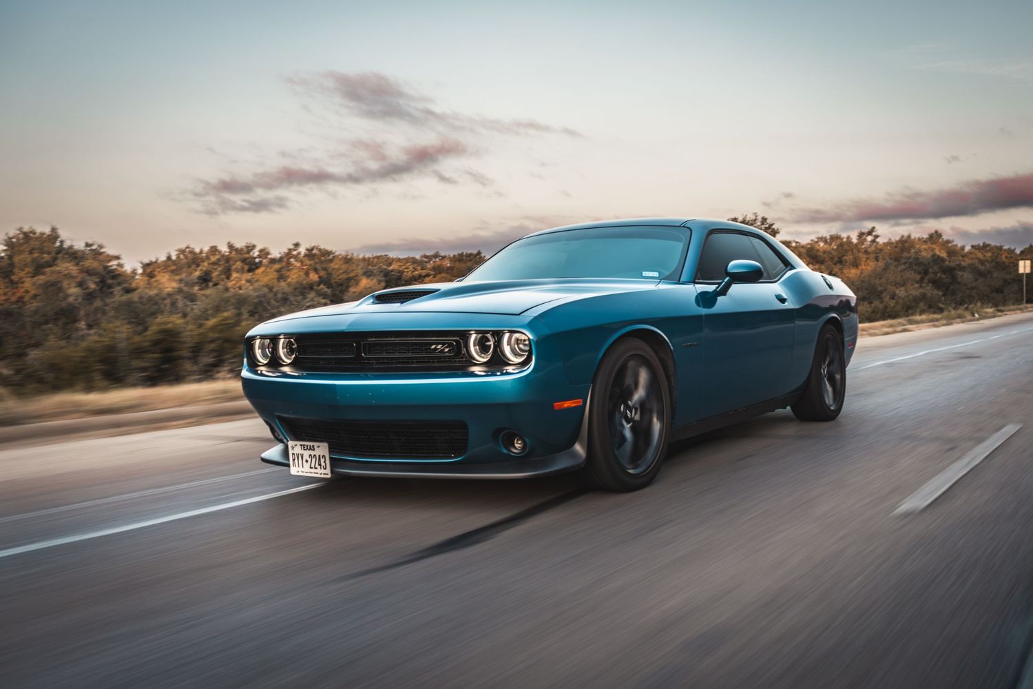 A blue Dodge Challenger muscle car speeds down an open road during sunset.