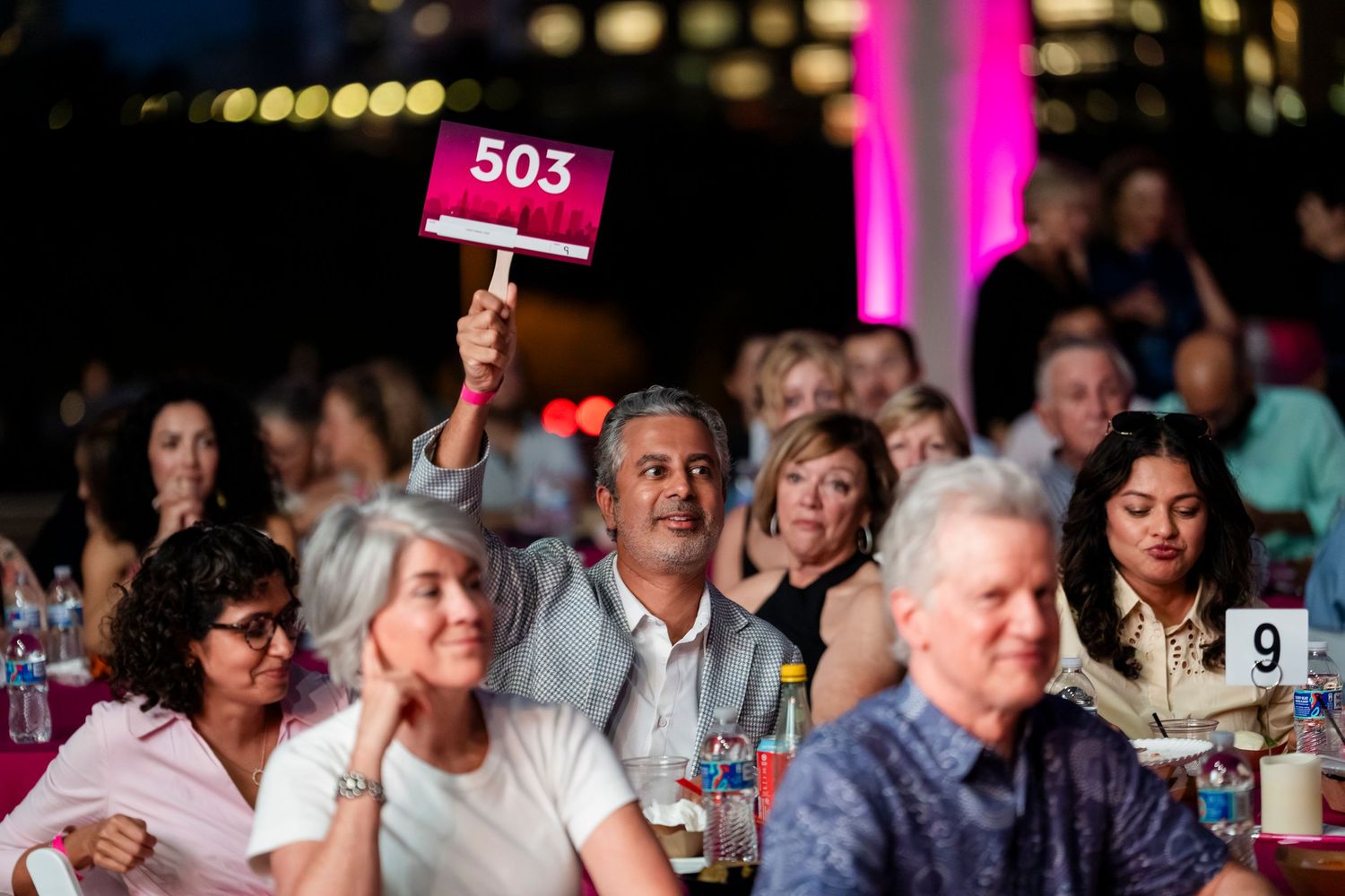 A large audience of people seated at tables during an indoor event with pink lighting and numbered table markers.