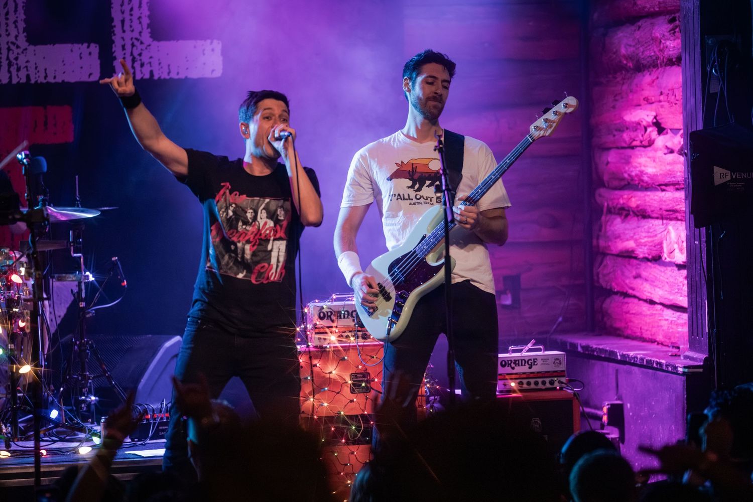 Musicians perform on stage with purple lighting and brick wall backdrop during live concert performance.