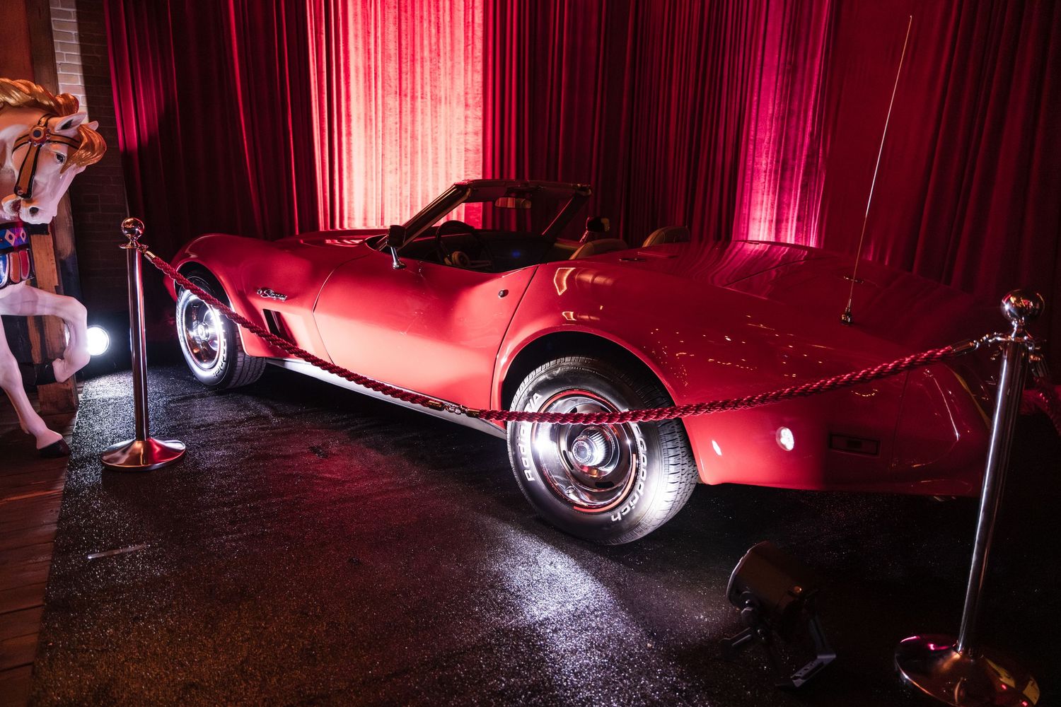 Red spotlights illuminate a classic car in dramatic low lighting at a nighttime auto show display.