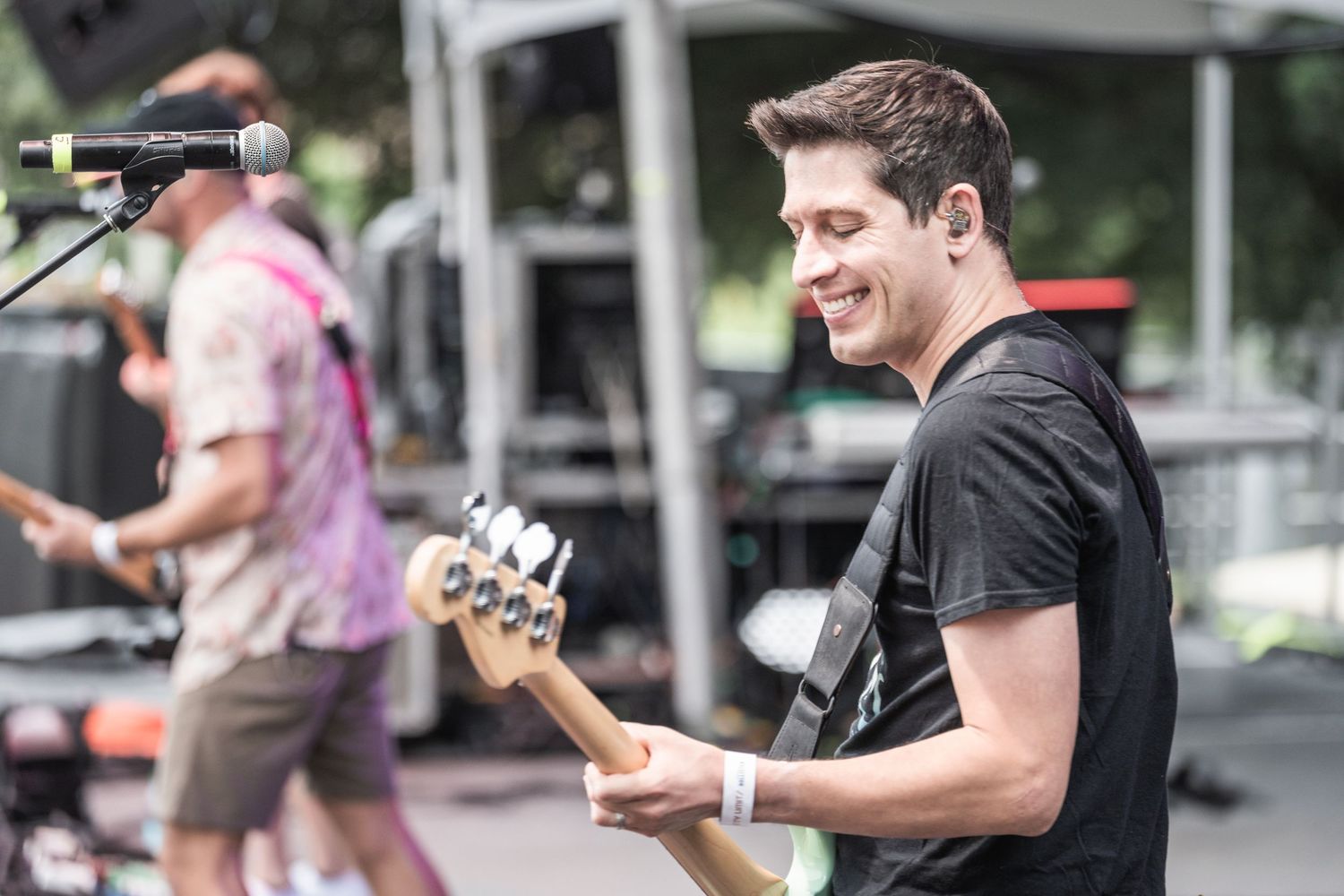 A person in a black t-shirt smiles while holding a mobile phone outdoors.