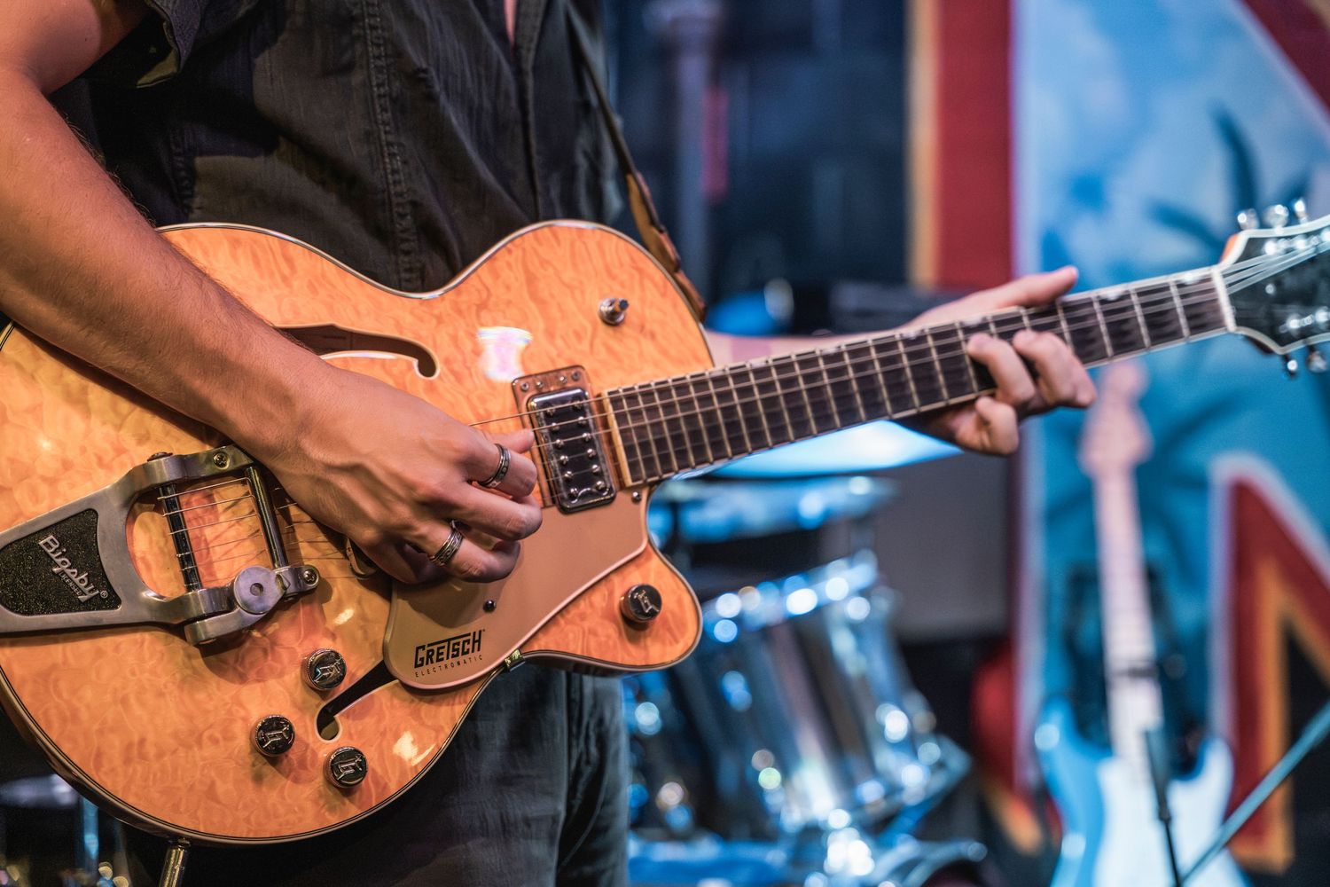 Close-up of hands playing a natural wood-finished hollow body electric guitar on stage with drum kit in background.