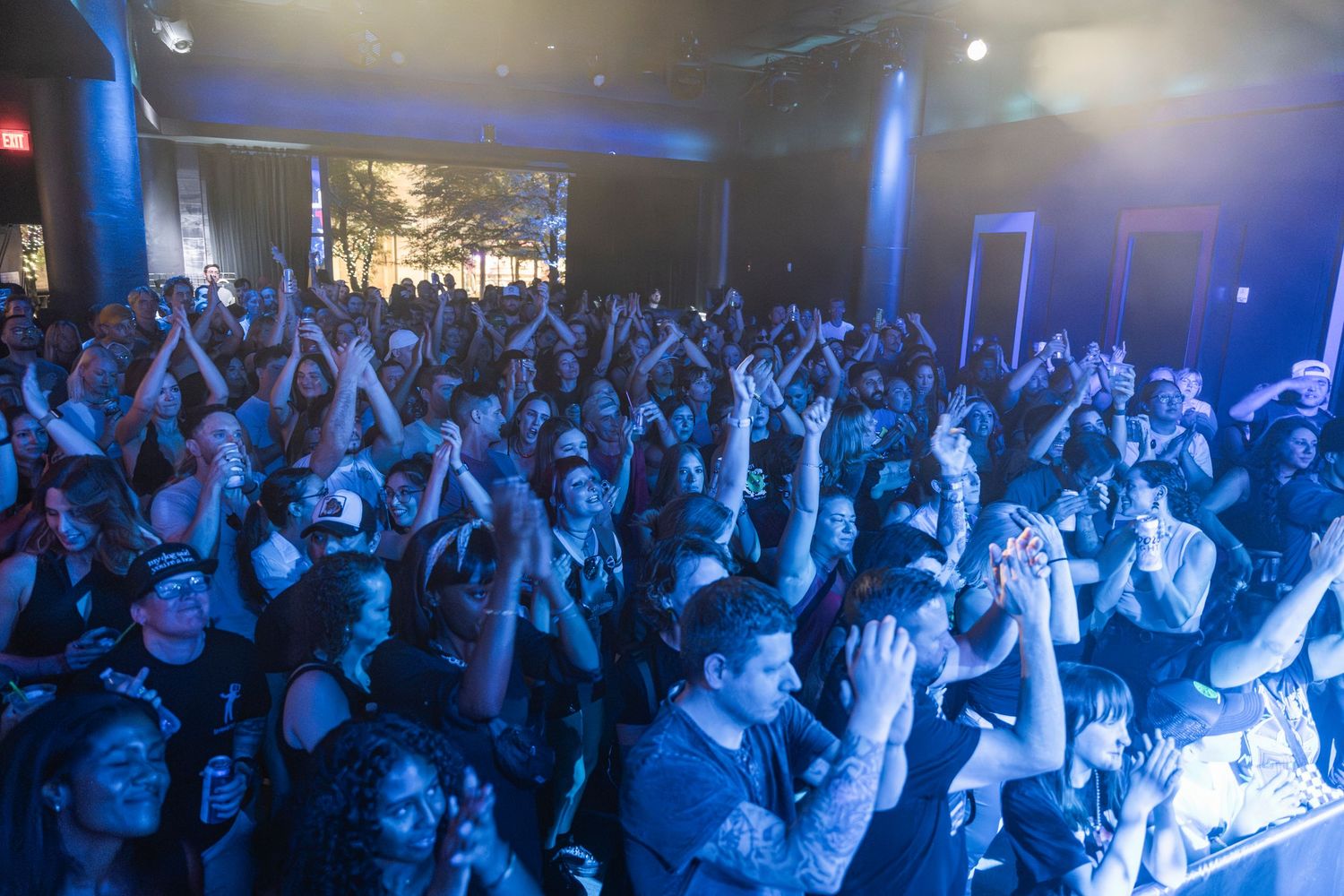 A large crowd of concert attendees with raised arms illuminated by blue stage lighting during a live performance.
