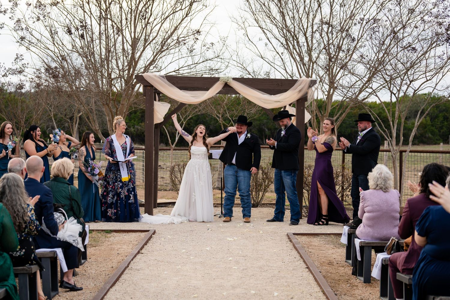 Bride and groom celebrate at outdoor rustic wedding ceremony under wooden arch with burlap decor, guests seated along aisle.